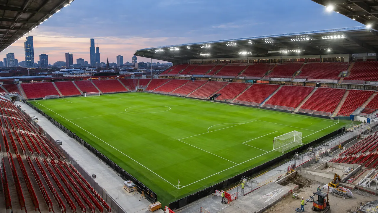 Newly rebuilt football stadium with red seats and a green pitch under floodlights, with finishing works at one corner